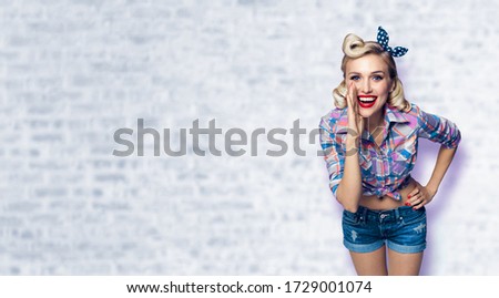 Beautiful happy excited woman holding hand near open mouth. Girl dressed in pin up. Blond model at retro fashion vintage concept, white brick wall background. Copy space area for sign or text.
