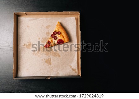 Closeup of the last whole slice of pepperoni pizza in a cardboard paper box on a dark background. View from above.