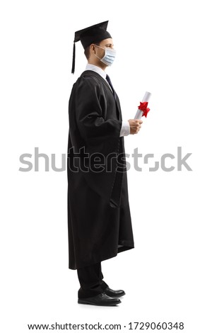 Full length profile shot of a male student in a graduation gown wearing a protective face mask and holding a diploma isolated on white background