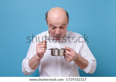 Senior funny caucasain man looking into a small empty pan while standing over blue background.