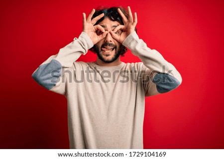 Young handsome man with beard wearing casual sweater standing over red background doing ok gesture like binoculars sticking tongue out, eyes looking through fingers. Crazy expression.