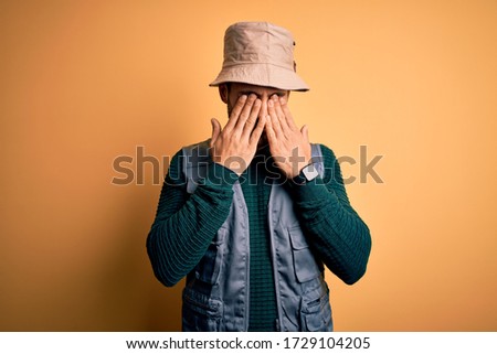 Handsome tourist man with beard on vacation wearing explorer hat over yellow background rubbing eyes for fatigue and headache, sleepy and tired expression. Vision problem