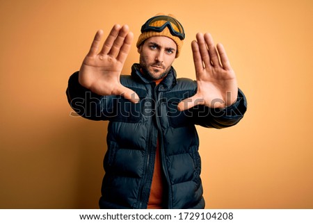 Young handsome skier man with beard wearing snow sportswear and ski goggles doing frame using hands palms and fingers, camera perspective