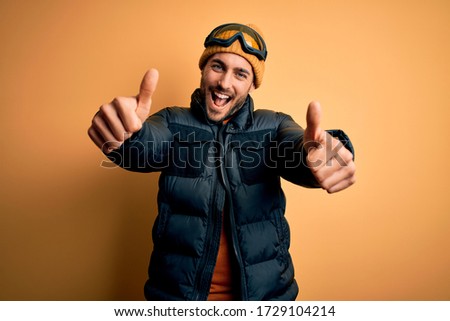 Young handsome skier man with beard wearing snow sportswear and ski goggles approving doing positive gesture with hand, thumbs up smiling and happy for success. Winner gesture.