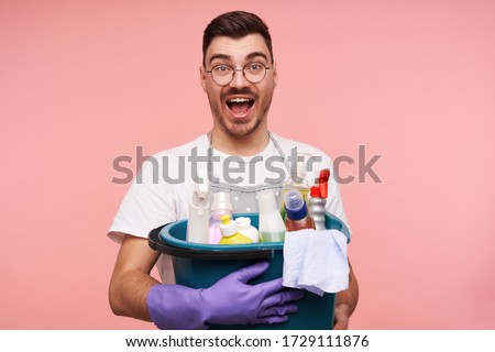 Overjoyed young handsome short brunette male in glasses dressed in apron and white t-shirt keeping busket with household chemicals and looking excitedly at camera with eyes and mouth opened