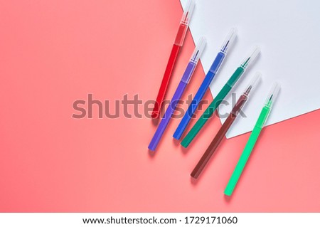 Multi colored felt pens and white blank paper sheet for drawing lies on pink desk in kindergarten. Сoncept of parenting and education of children. Copy space. Top view