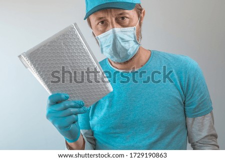 Delivery man posing with parcel package in protective clothing wearing baseball cap, protective mask and gloves during viral infection pandemic outbreak