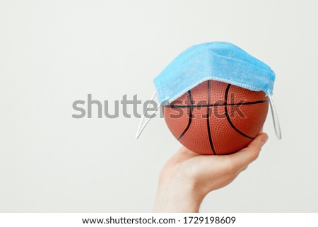 Basketball ball with medical mask on hand of man on light background with copy space. Coronavirus suspends sports world.