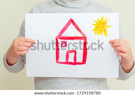 Closeup of child is holding picture of red house with sun at elementary school. Painting concept.