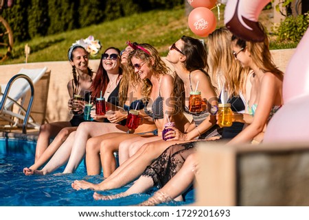 Group of girlfriends at a poolside summer party sitting at the edge of a swimming pool drinking and having fun.