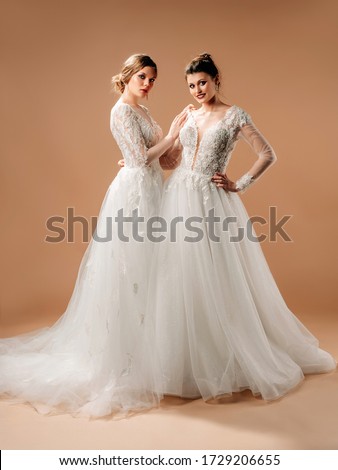 Wedding gown fashion. Beautiful happy brides with smile and long white wedding dresses posing in beige studio. Full-length portrait with neutral background. Wedding inspiration.