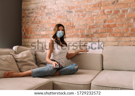 woman in medical mask holding remote controller and watching tv on sofa with stay at home sign during self isolation