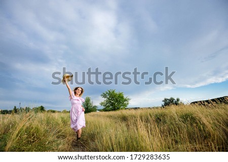 A cheerful young woman runs along the road, raising her hand with a straw hat, against the background of a cloudy sky. The concept of outdoor recreation in the countryside.