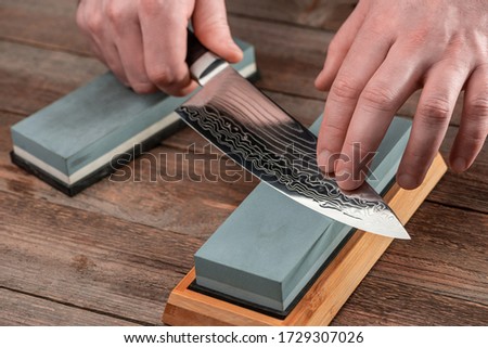Man sharpens a Gyuto knife using a whetstone on a rustic wooden table. Japanese knife with Damascus steel. Royalty-Free Stock Photo #1729307026