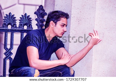 Lonely Asian American teenager thinking in New York City, wearing black T shirt, fashionable jeans, squatting against column by black metal railing outside office building. Color filtered effect.