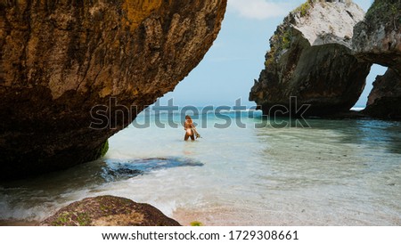 Blond long-haired surfer girl walking to the ocean  between the rocks holding her surfboard, female surfer holding copy-space surfboard