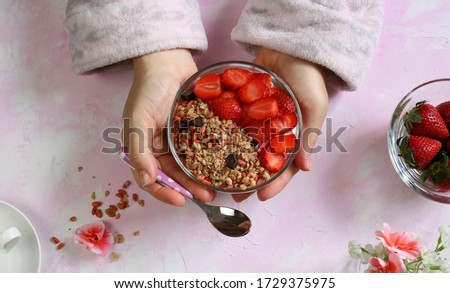Breakfast concept. Woman hands holds a bowl with strawberries, cereals and yogurt on white background with shades of pink. Top view.