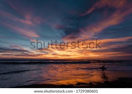 Bali, Indonesia. Dramatic landscape with sunset sky with bright orange clouds. Skyline, sky reflection on the sea water. Silhouette of a surfer walking along the shore with a board. 