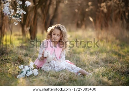 blond-haired girl of 7 years old, sitting in the field. holding a toy in his hands. dressed in a long lace white dress and a pink vest. field in the background