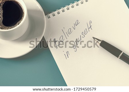 notepad with the inscription of a ballpoint pen believe in yourself. A cup of black coffee and a notebook with a pen on the blue table. Morning coffee
