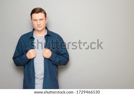 Studio portrait of focused blond mature man wearing casual blue shirt, with serious expression on face, attentively listening or waiting somebody, standing over gray background, copy space on right