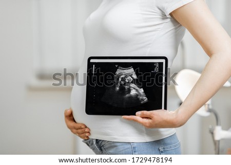 Pregnant woman holding digital tablet with an ultrasound scan of her unborn baby, standing in medical office, close-up view