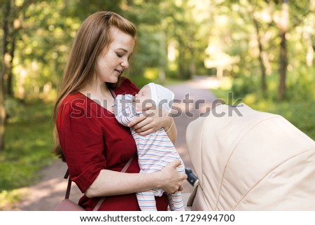 Young mother in the red dress hugs and caresses a two-month-old newborn baby in the summer green park. Walking with the baby, recovery after childbirth