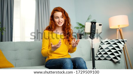 Portrait of young female blogger sitting on couch and talking in front of smartphone webcam. Girl coaching for her vlog online from home. Attractive happy woman coach talk for videoblog. Royalty-Free Stock Photo #1729509100