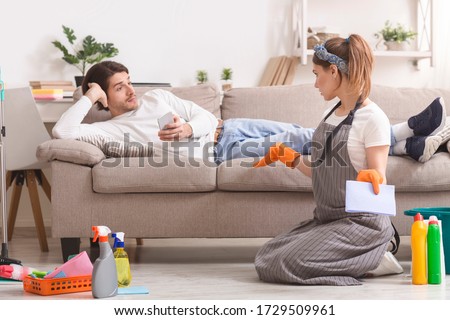 Frustrated Woman Cleaning Flat And Arguing With Her Lazy Husband Lying On Couch, Not Sharing Household Chores Royalty-Free Stock Photo #1729509961