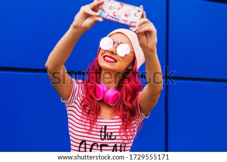Young, cute, funny girl, hipster with pink hair makes a selfie on a cell phone, listens to music, has fun, laughs, beautiful smile, hat, sunglasses, summer style