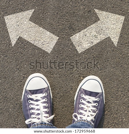 The person wearing sneakers stands on the asphalt road with two arrows, Concept of decision making.