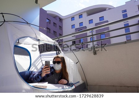 A girl in checked shirt, medical mask is lying in tourist tent at campsite and looks at phone.Concept of tourism after coronavirus and quarantine. Funny tourist at home on balcony, traveling Royalty-Free Stock Photo #1729628839