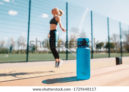 Ecologically clean water concept. A bottle of water for sports. Against the background of a bottle, a girl jumps on a rope. Sports ground. Blue colors. Street sport.