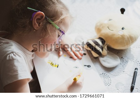 A little girl with glasses draws a portrait of a soft toy bee that lies in front of her. Idea for quarantining a child