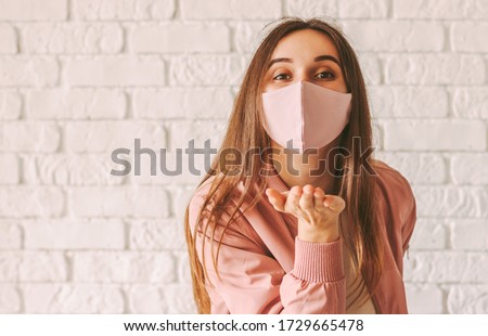 Portrait young happy millennial woman in trendy medical face mask blowing air kiss with hand. Beautiful stylish hipster girl in pink protective mask send blow kiss. Girl gesture blow kiss in face mask Royalty-Free Stock Photo #1729665478