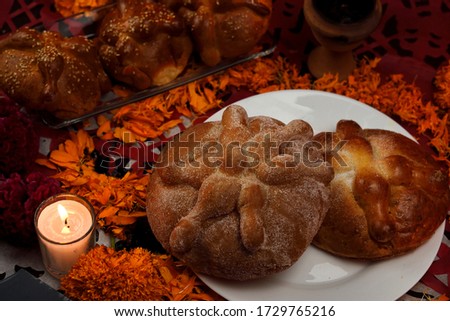 Traditional and sugary bread of the dead on a white glass plate, around it marigold flowers, lighted candles, diffuse background of colored confetti.