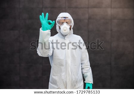 Portrait of man in sterile uniform and mask standing outdoors and showing okay sign. Surfaces are all sterilized from corona virus/ covid-19. Royalty-Free Stock Photo #1729785133