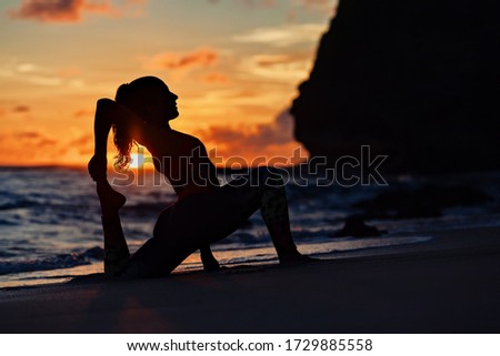 On sunset sea beach sporty young woman doing exercise, stretching to keep feet. Healthy lifestyle dark background. Outdoor sports activity at tropical island yoga spa retreat, family summer vacation.