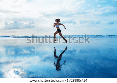 Barefoot young girl with slim body running along sea surf by water pool to keep fit and burning fat. Beach background with blue sky. Woman fitness, jogging sports activity on summer family vacation. Royalty-Free Stock Photo #1729885714