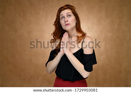 Model actress shows different emotions while talking on camera in the studio. Photo of a pretty caucasian young woman with minimal makeup in a black blouse with long red hair on a beige background.