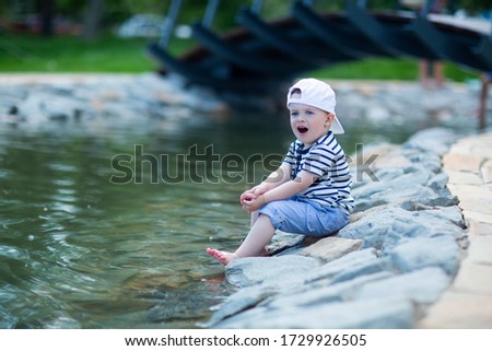the baby in a white cap sitting by the pond touches cold water with his leg and admires