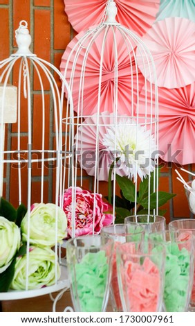
White metal cage with a white flower and another pink flower among the colorful decoration of a party.