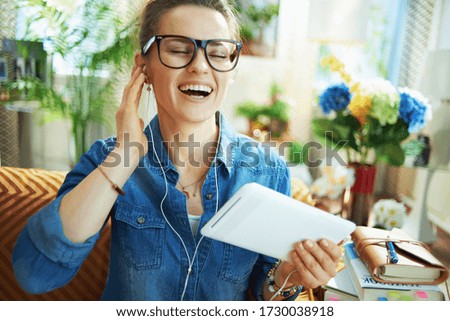 smiling stylish middle age woman in jeans shirt with white headphones and tablet PC study online in the modern living room in sunny day.
