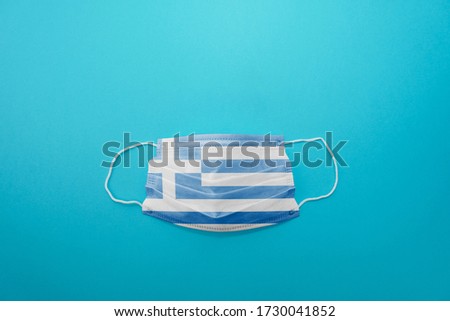 Disposable medical surgical face mask with Greece flag superimposed on it with blue background. Coronavirus (COVID-19) pandemic affects the country. Stay home, stay safe