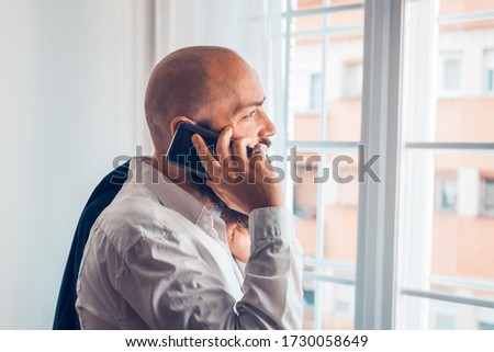 Bearded businessman in white shirt speaking on cellphone in office looking through window.
