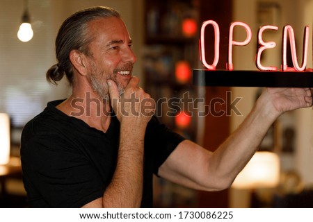 open for business concept, small business owner reopening premises after covid-19 virus pandemic, happy man looking at red neon open sign at a bar restaurant or cafe window after coronovirus lockdown