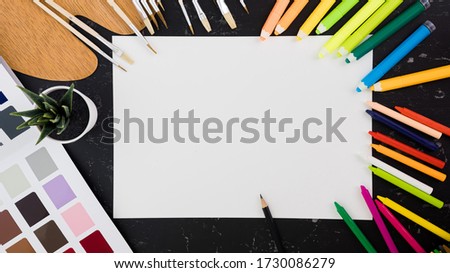 Top view of black desktop with piece of white paper and various colorful drawing tools.