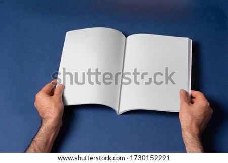 Male hands holding an opened book-catalog with blank pages on blue background, mock-up series template ready for your design, pages selection path included