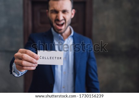 Selective focus of angry collector shouting and showing card with collector lettering in room