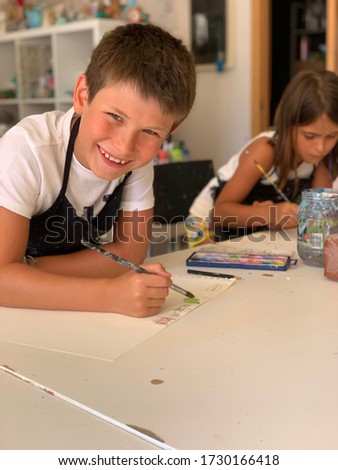 
Children in the drawing lesson paint. In the foreground, a close-up shows a smiling boy holding a brush in his hands. Image with selective focus y noise effects.